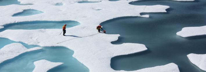 An aerial view of two people in orange coats and black pants standing on a piece of ice. One is close to the edge and bending over while picking up a canister. The edges of the ice scalloped and almost lace-like in appearance. The water visible ranges from light to deeper blues.
