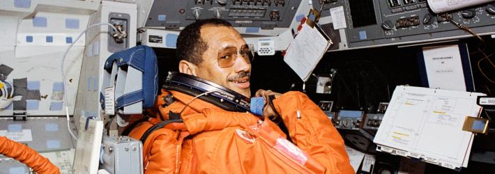 Astronaut Charles F. Bolden, a Black man, looks over his right shoulder and smiles at the camera. He is wearing an orange launch and entry suit and square tinted glasses without temples. He is sitting at the commander's station, which has many switches and dials, along with a notebook, a 3-ring binder, and various sheets of paper.