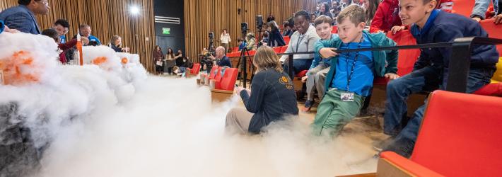 Fog rolls across the floor of an auditorium full of students and teachers. In the background at left are NASA astronauts Jessica Watkins and Bob Hines, along with students and teachers. To the right are rows of orange seats, where the audience sits, watching the experiment underway.