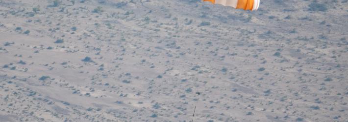 An orange and white parachute is visible at top right. It is attached to a training model of a capsule. The black and white cone-like capsule is quite small in this image. The pale ground below makes up the background of the image.