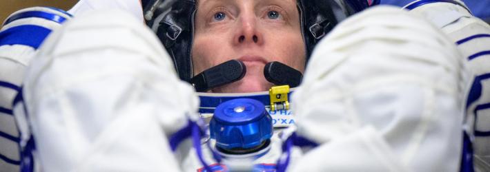 Astronaut Loral O'Hara looks upward while in a reclined position. Her feet are out of focus in the foreground. Her hand rests on the blue rim of her helmet. She wears a white and blue spacesuit.