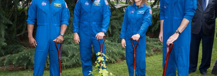 Four astronauts, three men and one woman (second from right), rest their hands on the handles of short shovels that are planted in dirt. They surround a small tree sapling that has bright green leaves.