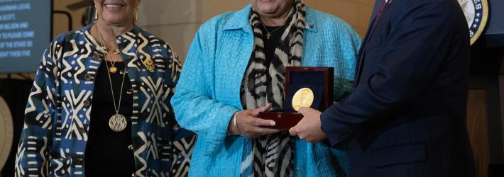 Two Black women and a white man face slightly left as they pose for a photo. The man and the woman in the middle hold a box containing a Congressional Gold Medal.