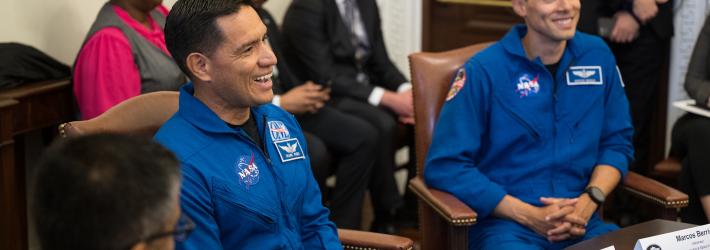 Two Hispanic men in blue jumpsuits with NASA patches on them sit at a table, smiling at people off screen. There are people of different ethnicities in the background.