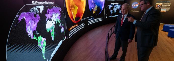 Two men in suits stand behind a short curved barrier. They look at a large curved screen that spans the room. The screen shows information, maps, and other graphics that relay information about carbon dioxide. The man on the right is pointing at the display.