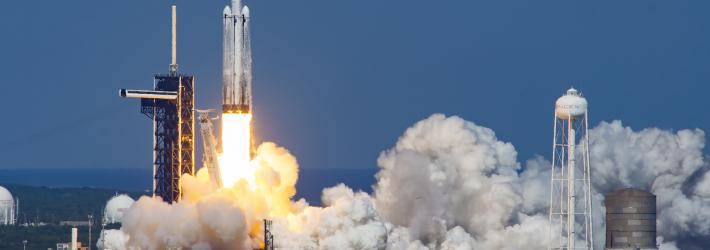 A white rocket lifts off from the launch pad, spewing billowing clouds of white vapor horizontally along the ground. A relatively short column of flames erupts from below the rocket just after liftoff. The early afternoon sky and the water in the foreground are similar, vibrant shades of blue.