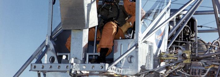 A man in an orange jumpsuit and white helmet sits in the pilot’s seat on a metal vehicle, the Lunar Landing Research Vehicle, or LLRV. The LLRV has a lot of metal tubing everywhere, which made people compare it to a metal bedframe.