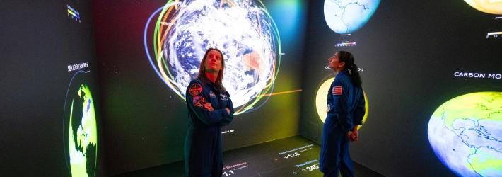 Two women in blue jumpsuits stand in a room with black walls and a black floor. The walls and floor have infographics, and data points on them related to Earth. The woman at left, NASA astronaut Loral O'Hara, looks up and to the side. The other woman, NASA astronaut Jasmin Moghbeli, faces the other way.