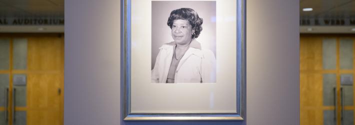 A black and white photo of Mary W. Jackson, an African American woman, is in a silver frame. The picture is displayed on a wall with a plaque underneath. Overhead light highlights the photograph.