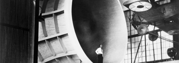 A man stands inside the entrance of a tunnel, looking up at a small propeller plane. The tunnel, viewed from the side, is like a cylinder with sides curving inward. The photo is in black and white.