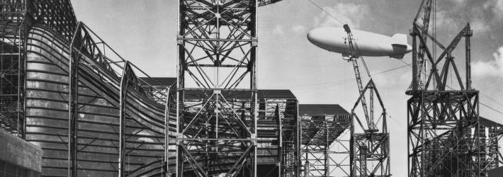 Black and white photo of a wind tunnel at Ames under construction. A Navy blimp is flying in the background.