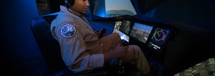 A man wearing a tan flight suit flies in a blue-lit flight simulator. Navigation guidance and other controls light up the simulator’s front dashboard.