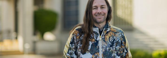 Garrett Sadler smiles softly outside in front of a marble building for his Faces of NASA portrait. His brown hair is long, past his shoulders, and he's wearing a zipped-up jacket with tropical plants, flowers, and tigers on it.