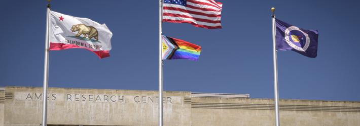 Three silver flagpoles are in front of a building with the words "Ames Research Center" on it. On the left pole is the California state flag. On the middle flagpole are, from top to bottom, the American flag and the Intersex Progress Pride flag. The Intersex Progress Pride flag has a purple circle on a yellow triangle, a five-stripe chevron with colors representing LGBTQI+ people of color and the transgender community, and rainbow stripes. Finally, on the right flagpole, is the NASA flag.
