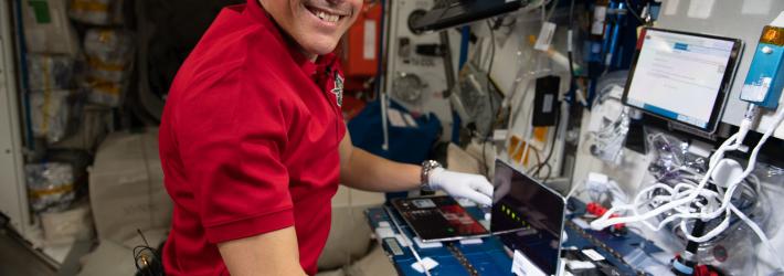 Bob Hines, wearing a red work polo and white gloves, smiles at the camera from his position at a workstation with many screens and wires aboard the International Space Station.