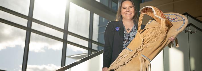 Tami, a woman of Native American heritage from the Delaware Nation, smiles standing on a staircase in a modern, sunlit building, holding a traditional handcrafted cradleboard adorned with intricate beadwork. She is wearing a paisley patterned shirt covered with a black sweater that has the NASA insignia on the right breast.