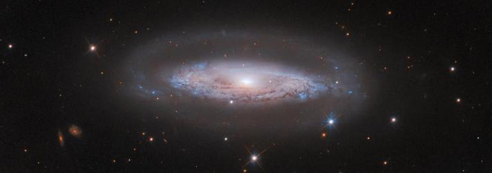 A spiral galaxy with a bright-white core, a glowing disk thick with swirling patterns of dark dust, and a faint halo around the disk. It is on a black background with a few small, distant galaxies and some foreground stars around it.