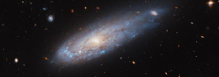 A spiral galaxy with a disk that glows visibly from the center. It has faint dust threaded through it. A spiral arm curves around the left edge of the disk and is noticeably denser with bright blue spots that hold hot and new stars. On the opposite side, the disk stretches out into a short tail where it covers a distant background galaxy. Other distant galaxies and some nearby stars are visible are also visible in this image.