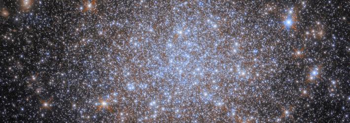 A cluster of stars. Most of the stars are very small and uniform in size, and they are notably bluish and cluster more densely together toward the center of the image. Some appear larger in the foreground. The stars give away to a dark background at the corners of the image.