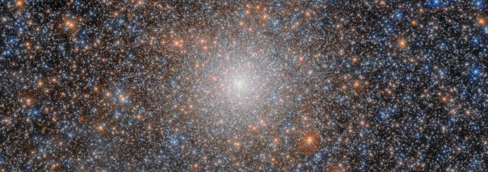 A globular cluster that looks like a very dense, ball-shaped collection of many shining stars in colors of white, yellow-orange, and blue. Some stars appear a bit larger and brighter than others, with the brightest having faint cross-shaped diffraction spikes. The cluster’s stars are scattered mostly uniformly, with their density increasing toward the cluster’s core where they merge into a strong, bright-white glow.
