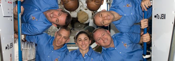 Seven astronauts pose for a photo on the International Space Station. They are arranged with their heads in a circle, with a few people upside down. They're all wearing blue polo shirts with an STS-129 logo on them.