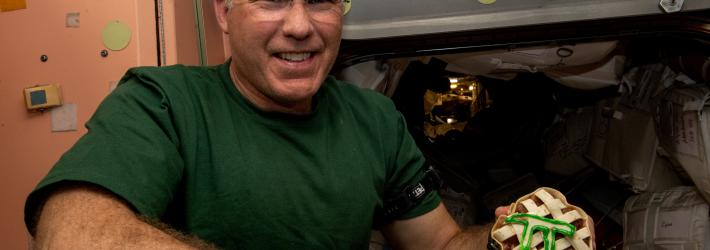 Stephen Bowen, wearing a green t-shirt and glasses, poses with a small latticed pie with a pi symbol outlined in green atop the lattice. Part of the International Space Station is visible behind him.