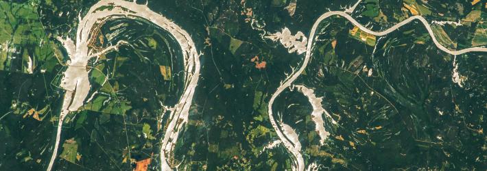 The silvery waters of the Alabama River meander across the image, contrasted against surrounding land in shades of green. At various points, the water extends outward from the borders of the river.