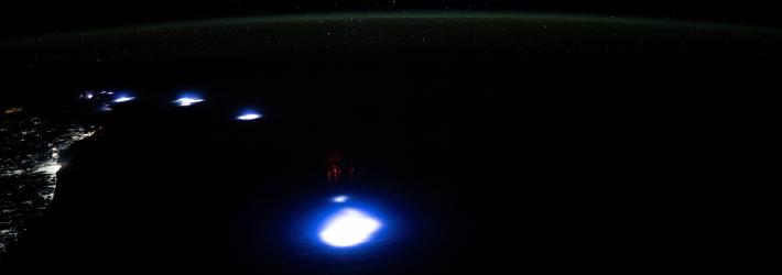 A line of bright blue lightning storms shines against a black background. Above the largest storm small wisps of red sprites stretch up. Along the left side, small yellow lights shine from an illuminated city.