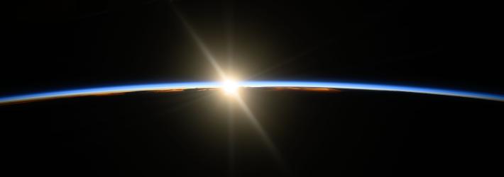 The beginning rays of an orbital sunrise illuminate Earth's atmosphere, a slight, blue arc across the center of the image. Earth is below, totally dark, matching the blackness of space above.