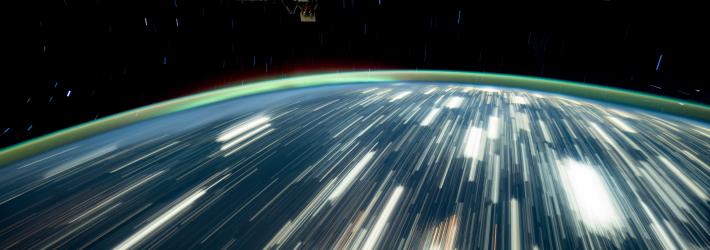 City lights on Earth appear as bright streaks in this long-exposure photo. Earth's atmosphere glows green on the horizon. Part of the International Space Station is visible at top right.