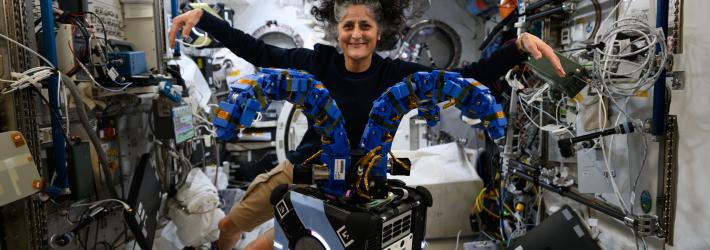 Astrobee, a robot that looks like a small black cube with two blue tentacle-like arms on top. Behind it, astronaut Suni Williams stretches her arms out to match Astrobee's arms.