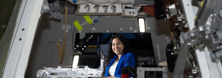 Zaida Hernandez, a hispanic woman with long wavy brown hair and dark brown eyes, smiles brightly at the camera while sitting inside a mock up of NASA's Orion Crew Capsule. She's wearing a blue blazer and flight hardware is seen all around her.