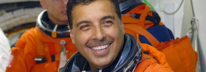 Astronaut José Hernández, a Hispanic man, smiles broadly at the camera while waiting to board space shuttle Discovery. He wears an orange escape suit that has an American flag patch on the left shoulder. Behind him are mission specialists Patrick Forrester (left) and Christer Fuglesang (back to camera).