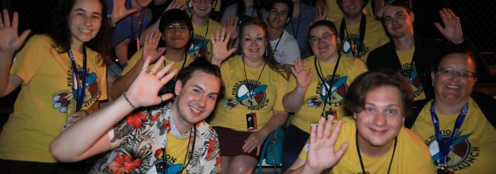 A group of people wearing yellow t-shirts wave at the camera as they sit outside at night. They are members or advisors of winning teams from the 14th First Nations Launch High-Power Rocket Competition. The competition comprises students from tribal colleges and universities, Native American-Serving Nontribal Institutions, and collegiate chapters of the American Indian Science and Engineering Society.