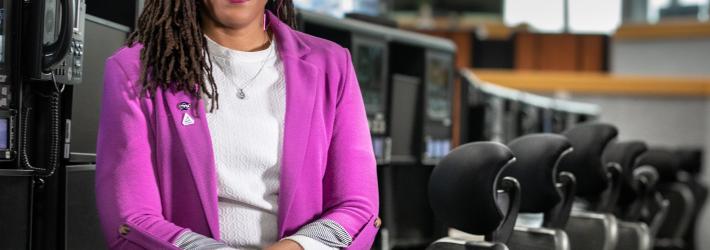 Ales-Cia Winsley smiles for the camera as she sits in front of a row of desks in Firing Room 1 in the Launch Control Center at NASA's Kennedy Space Center. She is wearing a bright magenta jacket with two pins (the NASA "meatball" insignia and the NASA Artemis logo) present on her right lapel, a white shirt, and striped slacks.