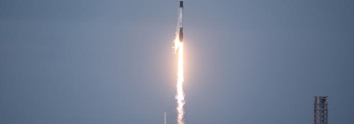 A black and white SpaceX Falcon 9 rocket launches upward through the sky, a bright white-yellow plume trailing behind it.