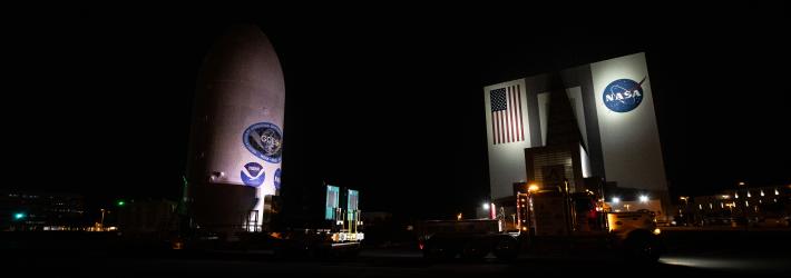 NOAA's GOES-U satellite (left) and NASA's Vehicle Assembly Building (right) are both lit up at night. GOES-U is a white cylindrical object and the VAB is a tall, square building.