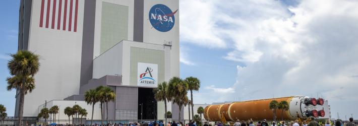 The Artemis II rocket core stage, a long orange cylinder with four red boosters) is on its side as it rolls into the Vehicle Assembly Building (VAB) at NASA's Kennedy Space Center while employees watch. The VAB is a tall rectangular building with the American flag and NASA "meatball" logo painted on its left and right side, respectively. There is also an Artemis logo on a lower portion of the building.