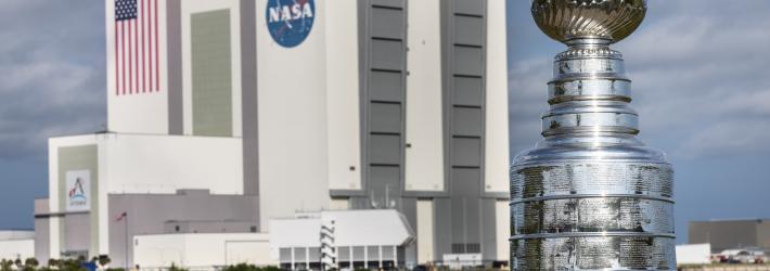 The silver Stanley Cup is in the foreground at right. The Stanley Cup looks like a large silver bowl set on top of a stacked column of thick silver layers. There are words engraved on the Cup. Behind it is a body of water and the Vehicle Assembly Building at NASA's Kennedy Space Center, a massive, boxy gray and white building.