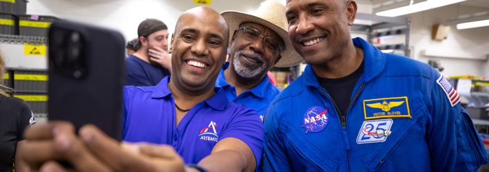 Three African American men - two Kennedy Space Center employees wearing blue polo shirts with an Artemis logo (left, middle back) and NASA astronaut Victor Glover (right) - pose for a photo taken with a cell phone.