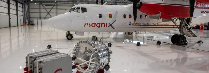 A four engine turboprop aircraft wrapped in a red and white livery with logos and names of each partner on the project sits under the lights inside an aircraft hangar. On the ground in front of the plane is an electric powertrain with an electric motor and battery pack that will soon be swapped out with one of the aircraft’s traditional engines to form a hybrid electric system.