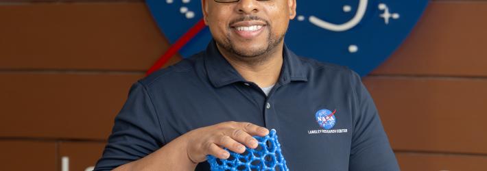 Acting Center Chief Technologist Dr. Phillip Williams holds up a model and smiles at the camera. A NASA meatball logo can be seen in the background.