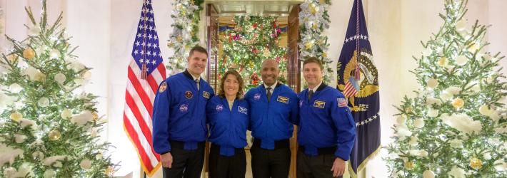 Four astronauts, wearing blue jackets and black pants, pose for a photo at the White House in front of decorations like trees, wreaths, and boughs. The astronauts, members of the Artemis II mission, have their arms around each other's shoulders. Behind them are two flags, and a seal high on the wall that reads "Seal of the President of the United States."