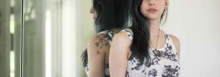 A woman with long, dark brown hair and subtle blue highlights stares head on at the camera wearing a white tank top with blue and purple roses on it.