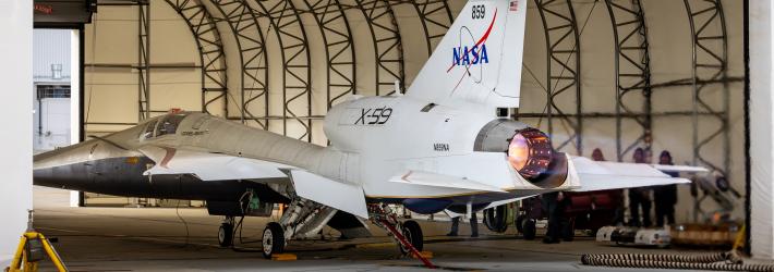 The nearly 100-foot-long X-59 sits partially inside a large run stall, with the rear of the aircraft and its engine extending outside the run stall’s open bay door. Flames are visible from the engine, producing additional thrust during full afterburner tests.