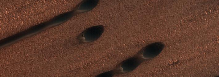 An aerial view of the reddish-brown surface of Mars taken by the Mars Reconnaissance Orbiter. Barchan dunes (circular dunes) appear here like oval divots in the ground. Linear dunes also appear.