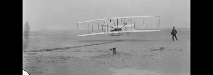 In this black and white photo, a white airplane with two sets of stacked wings with wires connecting them flies low to the ground. A man, Wilbur Wright, stands on the right with his back to the camera.