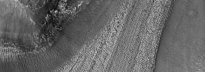 A black and white image of a portion of Mars' surface from above. Ridges stretch from top right to the bottom, the result of ice moving over possibly thousands of years. The glacier-like forms left behind have a mottled appearance.
