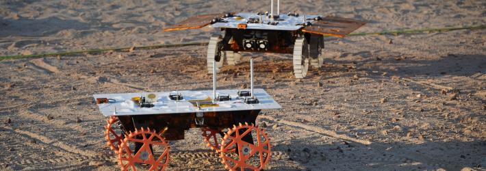 Two small rovers in a sandy yard. The rover in the back has solar panels fitted to it and white wheels. The one in the front has orange wheels and no solar panels.