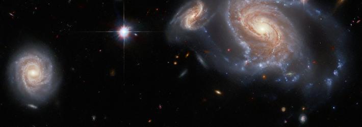Against the blackness of space, one very large spiral galaxy and three smaller spiral galaxies are prominently visible amidst the other colorful specks of light.